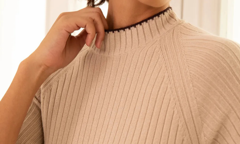 Close em look premium com tricot básico bege claro, destacando a gola alta canelada e os detalhes do ponto do suéter em textura macia e elegante.