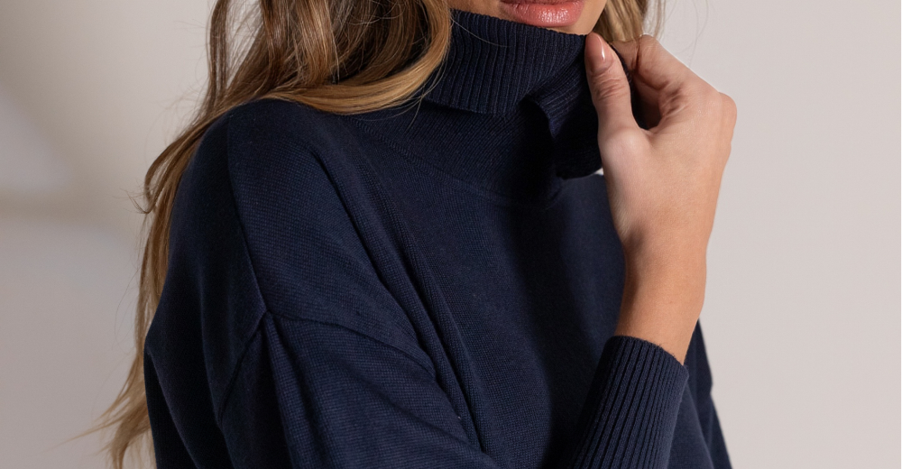 Mulher com suéter de gola alta azul marinho, segurando a gola com a mão, em close. Visual elegante e minimalista que inspira ideias de guarda-roupa cápsula para combinações versáteis.